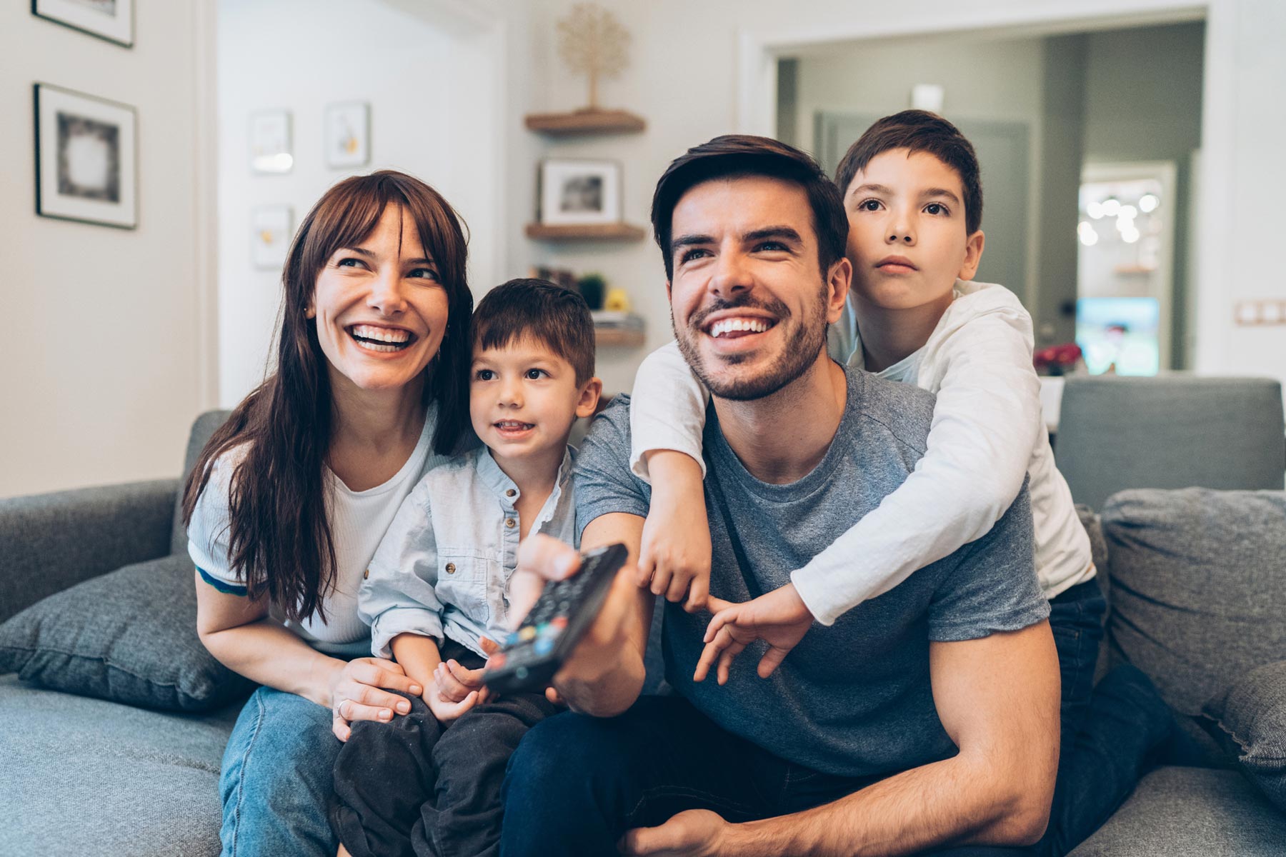 Family sits together on a couch smiling and pointing a remote control at a TV