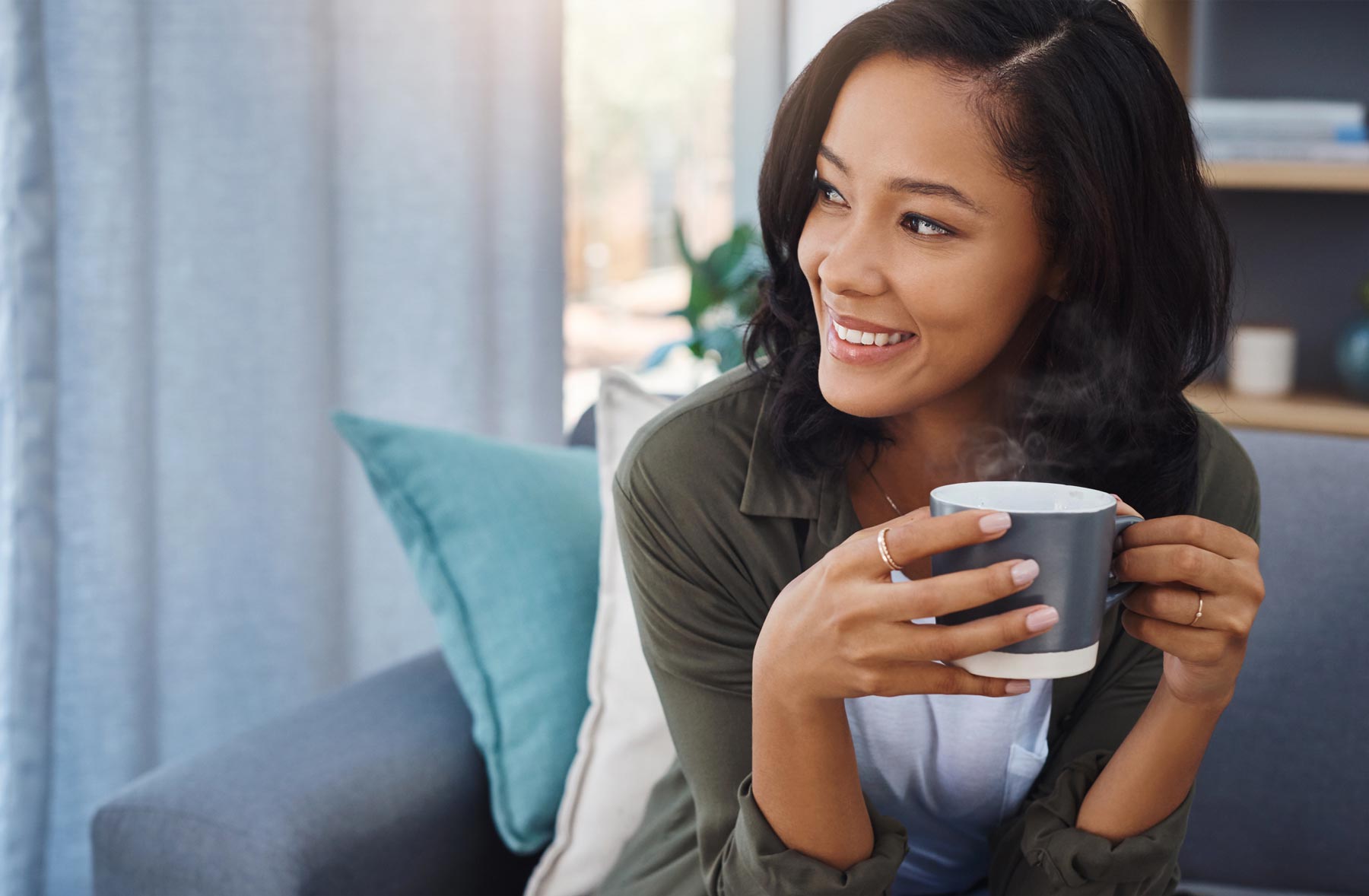 Woman smiling and looking away while holding a steaming mug on a living room couch