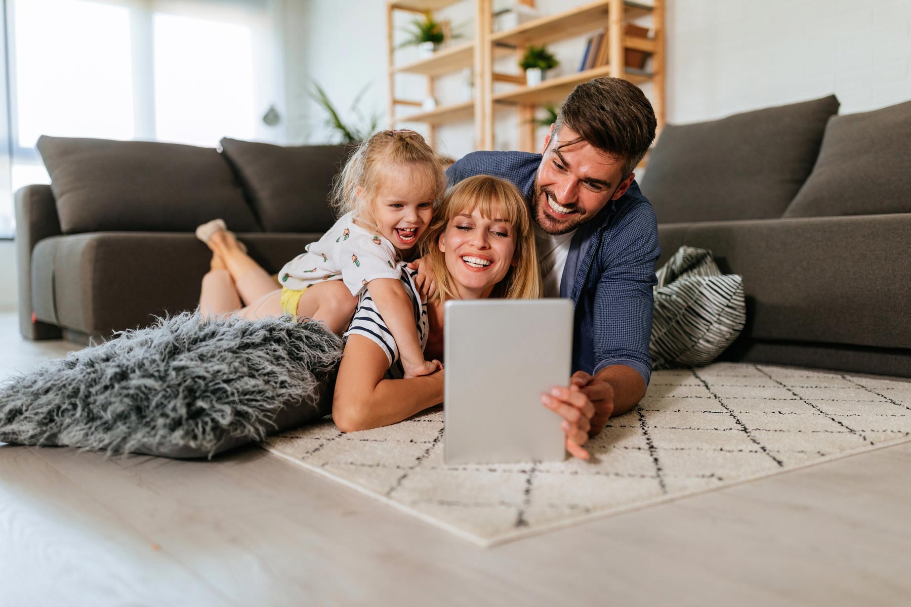 Parents and young daughter lie on a living room rug, smiling while looking at a tablet together