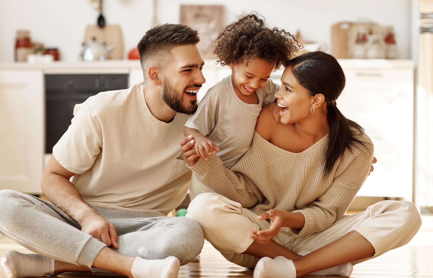 Parents and a young child sit on the floor laughing together in a bright kitchen