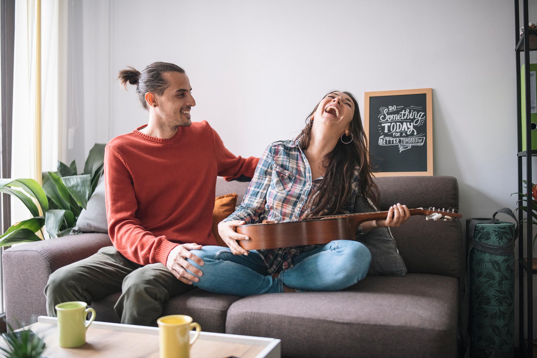 Man and woman laugh together on a sofa as she plays guitar in a bright living room