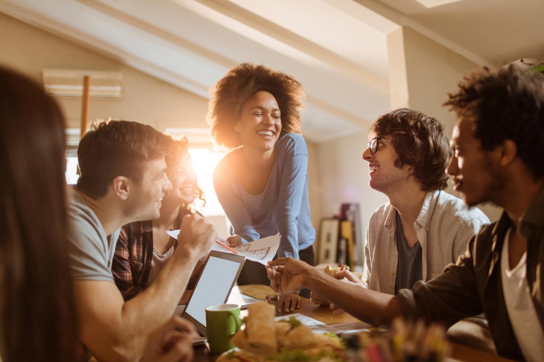 A diverse group of friends smile and chat around a table with a laptop, coffee, and sandwiches